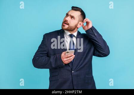 Uomo d'affari vestito ufficiale stile gratta testa tenendo smartphone in mano, contemplando l'aggiornamento del software, scegliendo tariffe adatte. Studio interno girato isolato su sfondo blu. Foto Stock