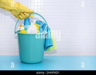 Una mano in guanto giallo tiene il secchio con detergenti e accessori per la pulizia, stracci guanti spazzole in spugna. Foto Stock