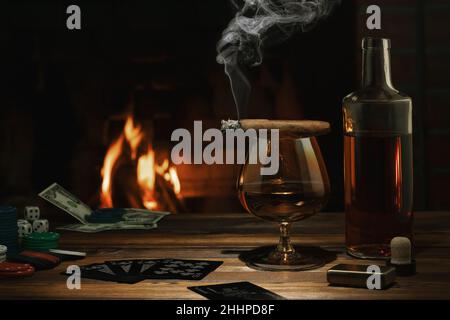 Bicchiere di cognac, sigaro, bottiglia, gioco di carte e fiches sul tavolo vicino al camino in fiamme. Riposo, piacere, buona fortuna e dolce vita concetto. Foto Stock
