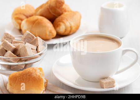 Caffè con latte in una tazza bianca, panini e zucchero di canna su un tavolo di legno bianco. Foto Stock