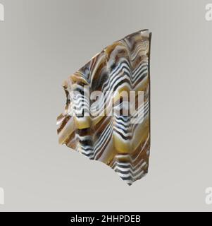 Mosaico di vetro dentato frammento di ciotola fine 1st secolo a.C.–inizio 1st secolo d.C. frammento di corpo romano.marrone di miele traslucido, blu di cobalto e bianco opaco.collo leggermente soprastato; Lato diritto rastremato verso il basso e curvato in basso. Motivo a mosaico a nastro formato da sezioni di una canna singola in marrone macinato con fili ondulati irregolari bianchi e blu in linee parallele; all'esterno, due ampie nervature verticali, abbastanza distanziate, con sommità appiattita e bordi esterni arrotondati.interno lucido; vaiolatura di bolle superficiali all'interno; sbucciature e macchie di cremoso iridescente agenti atmosferici all'esterno Foto Stock