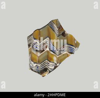 Mosaico di vetro dentato ciotola frammento fine 1st secolo a.C.–inizio 1st secolo d.C. corpo romano frammento.marrone di miele traslucido, parzialmente mescolato con bianco e apparente giallo opaco, blu, E bianco opaco. Lato quasi diritto, rastremantesi verso il basso. Disegno a mosaico a nastro formato da lunghezze di una canna singola in una terra marrone con bande a zig-zag di fili bianchi e blu in linee parallele e bande piane in giallo; Sull'esterno, parti di tre nervature verticali, con parti superiori appiattite e bordi esterni arrotondati.esterni lucidati; vaiolatura di bolle di superficie e di alcuni trucioli stagionato sull'esterno; sverdimento e cremoso i Foto Stock