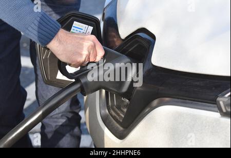 La mano e il braccio di un uomo che tiene un cavo del veicolo elettrico mentre collega la sua auto elettrica a un caricatore per auto EV presso una stazione di servizio autostradale Foto Stock