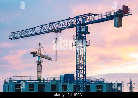 Cantiere con gru, nuovi edifici o case in costruzione con operai con casco al lavoro e cielo colorato sullo sfondo Foto Stock