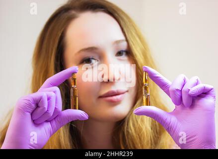 Un giovane cosmetologo sorridente in guanti di gomma viola sta tenendo fiale di siero di sollevamento. Un concentrato in fiale per correggere le rughe, restori Foto Stock