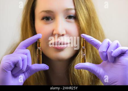 Un giovane cosmetologo sorridente in guanti di gomma viola sta tenendo fiale di siero di sollevamento. Un concentrato in fiale per correggere le rughe, restori Foto Stock