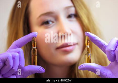 Un giovane cosmetologo sorridente in guanti di gomma viola sta tenendo fiale di siero di sollevamento. Un concentrato in fiale per correggere le rughe, restori Foto Stock