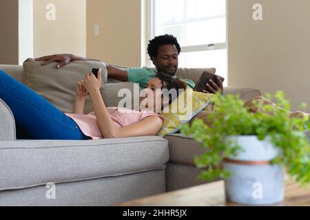 Giovane coppia multirazziale che usa smartphone e tablet pc mentre si rilassa sul divano a casa. Persone, convivenza e tecnologia concetto, inalterato. Foto Stock