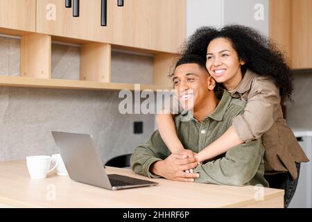 Simpatica coppia multirazziale che si gode il tempo libero a casa con un portatile. Donna e uomo felici in abbraccio guardando lo schermo e ridendo, guardando video, videochiamate con gli amici Foto Stock