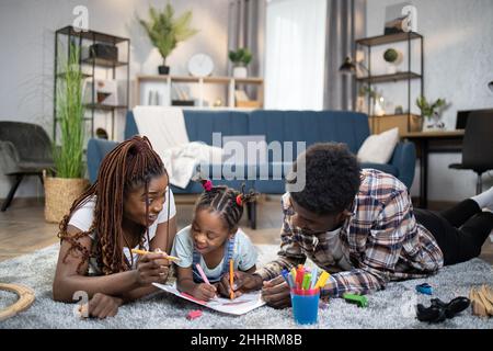 La figlia gioiosa e i suoi giovani genitori che si preoccupano di usare pennarello colorato per disegnare in album. Famiglia afroamericana di tre persone che si trova sul pavimento e gode di un processo creativo. Foto Stock