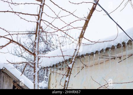 Le ghiacciole pendono dal tetto di una casa di villaggio in una giornata invernale. Scioglimento della neve. Foto Stock