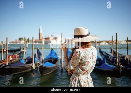 Vista da dietro elegante viaggiatore solista donna in abito floreale con cappello sul terrapieno a Venezia, Italia. Foto Stock