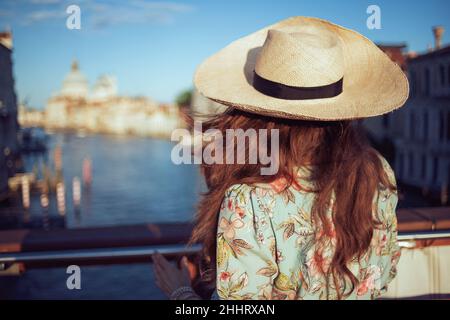 Vista da dietro l'elegante viaggiatore solista donna in abito floreale con visita del cappello sul ponte dell'Accademia a Venezia, Italia. Foto Stock