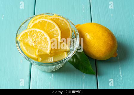 Fette di limone crude in un recipiente di vetro e una frutta intera su un tavolo di legno turchese. Agrumi freschi di colore giallo tagliato per concetti di vitamina C, antiossidante. Foto Stock