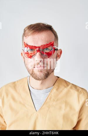 Ritratto da vicino di un medico o infermiera superman maschio. Ritratto di un bel medico caucasico maschio indossare maschera di supereroe viso rosso, occhiali e beige uniforme medica. Foto verticale di alta qualità Foto Stock