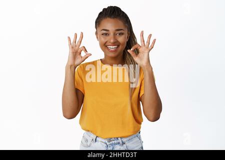 Happy Black ragazza sembra allegro, mostra OK segno, approvando, raccomandare prodotto, in piedi su sfondo bianco Foto Stock