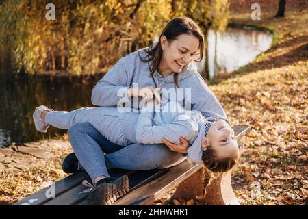 Madre seduta su panchina nel parco autunnale che tiene in braccio sua figlia con cui ha grande divertimento nei fine settimana. Ritratto di madre e figlia che hanno Foto Stock