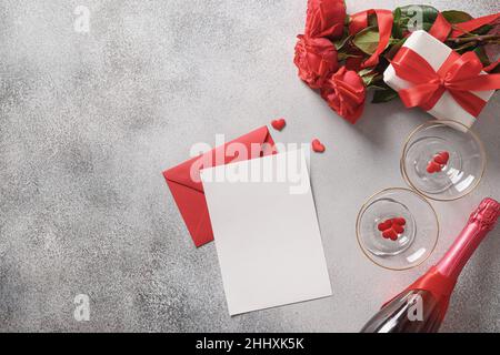 Biglietto di auguri per San Valentino con spazio vuoto per lettera d'amore, regalo, rose rosse, champagne e bicchieri da vino su sfondo grigio. Vista dall'alto. Copia Foto Stock