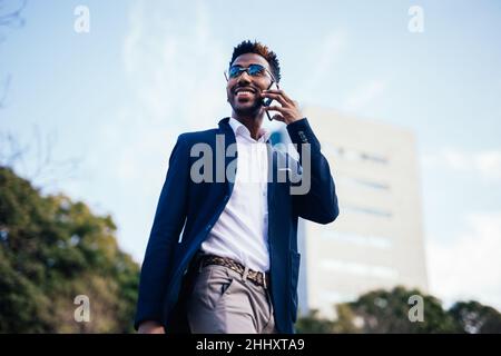 Primo piano di felice nero ombre imprenditore a piedi lungo la strada, mentre utilizzando il suo telefono fare affari Foto Stock