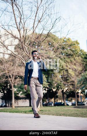 Grandangolo sorridente uomo nero imprenditore a piedi attraverso un parco mentre usando il suo telefono fare affari Foto Stock