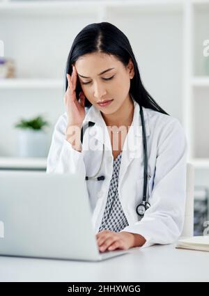 Quando il medico ha bisogno di un medico. Scatto di un medico giovane che osserva stressato mentre usando un laptop alla sua scrivania. Foto Stock