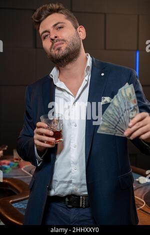 Giocatore di poker di successo con banconote e bicchiere di bevanda nel casinò Foto Stock