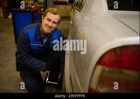 Bell'uomo caucasico, meccanico automatico professionale cambia manualmente la ruota in auto, svitando i dadi e i bulloni con la chiave, sorridendo al ° Foto Stock