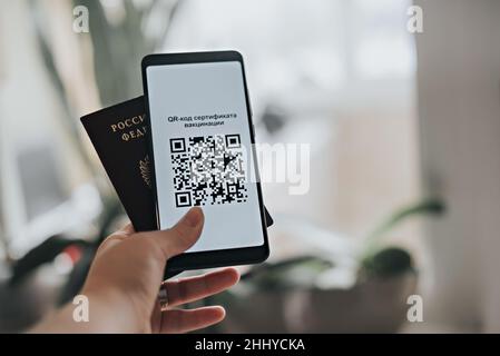 Mano della donna con passaporto russo e telefono con testo in russo - Codice QR del certificato di vaccinazione. Foto Stock