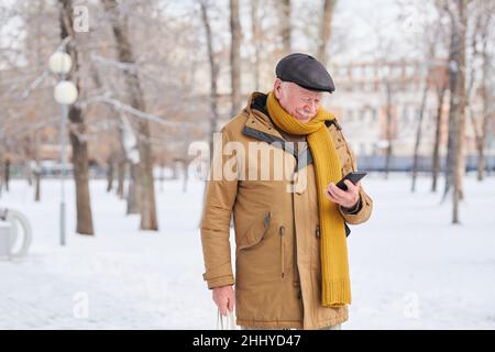 Uomo anziano in casual winterwear scorrimento in smartphone o lettura messaggio mentre si trova in piedi nel parco il giorno d'inverno Foto Stock