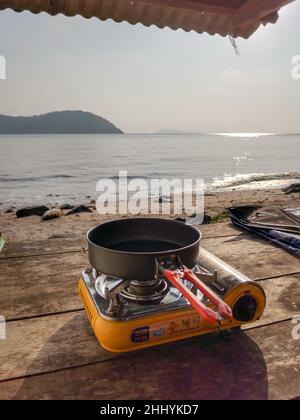 Stufa turistica portatile a gas e padella da cucina su tavolo di legno su sfondo spiaggia sabbiosa con luce del mattino. Attrezzatura da campeggio all'aperto. Campeggio più vitale Foto Stock