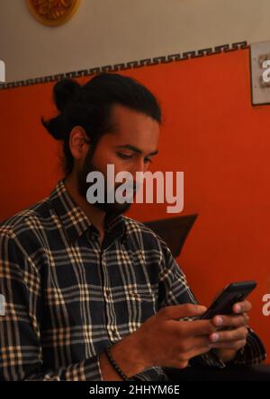 Vista laterale di un bel ragazzo bearded giovane con i capelli bun, usando il suo telefono mentre si siede al coperto Foto Stock
