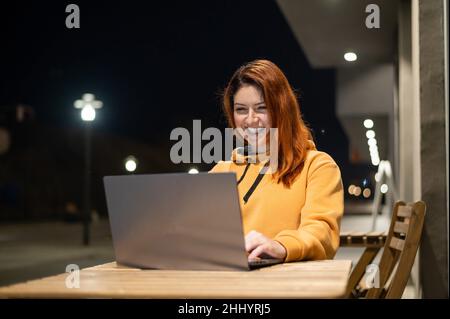 Una donna lavora a distanza a un computer portatile in un bar estivo a tarda sera. Ragazza felice studiare mentre si siede su una strada vuota ad un tavolo di legno Foto Stock