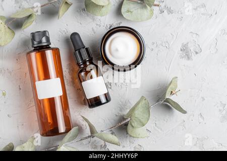 Bottiglie in vetro cosmetico con crema, toner e maschera facciale su fondo concreto con foglie di eucalipto secche. Il concetto di bellezza naturale cosmetici Foto Stock