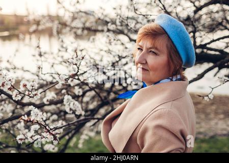 Giornata internazionale delle donne. Donna anziana di stile che cammina nel parco fiorito di primavera vicino al fiume ammirando il paesaggio naturale. Felice signora matura gode il tempo ou Foto Stock