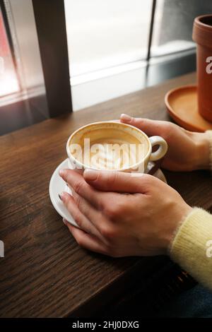 Donna mani in maglione giallo che tiene una tazza di caffè con latte art su sfondo di legno. Cappuccino. Pausa caffè. Concetto di hygge. Foto verticale Foto Stock