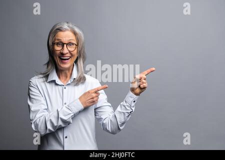 Donna d'affari positiva indossando abiti di stile ufficiale puntando dito lontano e prestando la vostra attenzione a spazio vuoto per la pubblicità mentre si guarda la macchina fotografica. Studio girato isolato su grigio Foto Stock