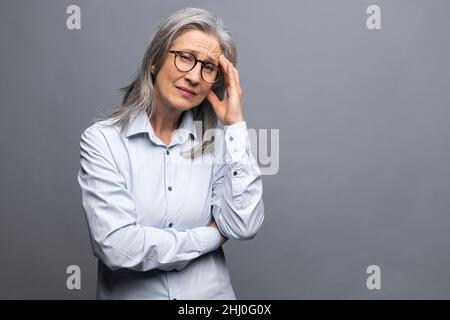 Una donna anziano si sente annoiato e stanco. Donna anziana con capelli grigi sconvolto che guarda la fotocamera e sente mal di testa e burnout isolato sopra la parete grigia Foto Stock