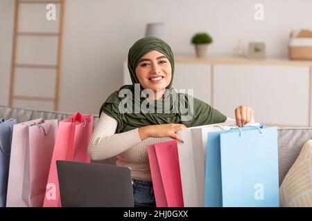 Donna araba felice in hijab seduta sul divano con laptop e borse luminoso shopper, acquisto di merci online a casa Foto Stock
