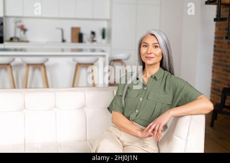 Headshot di anziano sognando asian etnico femmina con bel sorriso e guardando la macchina fotografica seduta contro sfocato interno casa sullo sfondo Foto Stock