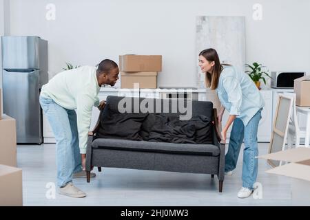felice coppia interrazziale che porta divano in soggiorno Foto Stock