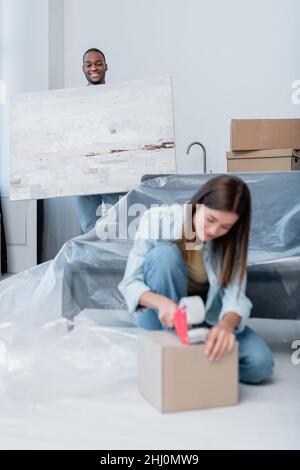 felice uomo afroamericano che tiene la pittura vicino alla scatola di imballaggio della giovane donna sfocata con nastro di regolazione Foto Stock