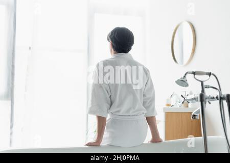 vista posteriore della brunette uomo in accappatoio bianco seduta su vasca Foto Stock