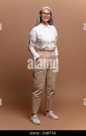Affascinante donna anziana sorridente toothy. Vista a tutta lunghezza dell'elegante e spensierata signora moderna dai capelli grigi che si posa alla telecamera isolata su sfondo beige Foto Stock
