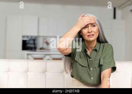 Donna preoccupata dai capelli grigi che tiene la fronte con mano seduta sul divano a casa, donna asiatica guarda via con disperazione, ha problemi o dimenticato qualcosa di importante, sente mal di testa Foto Stock