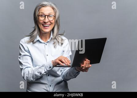 Donna anziana moderna in abbigliamento formale utilizzando computer portatile isolato in grigio. Ritratto di una donna matura dipendente ufficio utilizzando la tecnologia online per fare affari, computer app per la contabilità Foto Stock