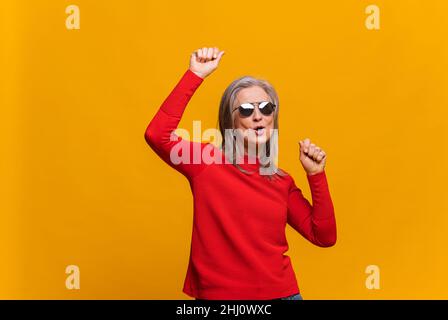 Concetto di umore di vacanza. Gioiosa e allegra donna anziana dai capelli grigi che alza le braccia e balla. Donna matura che indossa occhiali da sole celebrando qualcosa isolato sul giallo Foto Stock