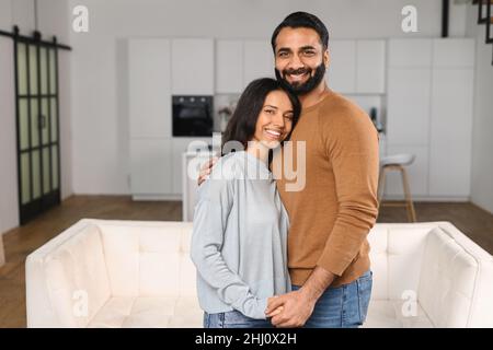 Primo piano di felice coppia interrazziale in posa su sfondo sfocato cucina. Felice proprietari di nuovo piatto sorridente e guardando la fotocamera. Giovane donna che abbraccia il suo bel marito Foto Stock
