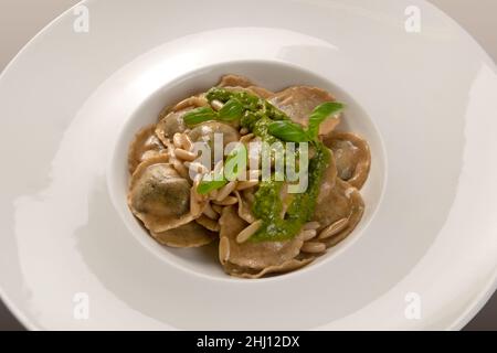 Ravioli rotondi (tortelli) di farina integrale con pinoli e pesto genovese e foglie di basilico in piatto bianco, da vicino Foto Stock