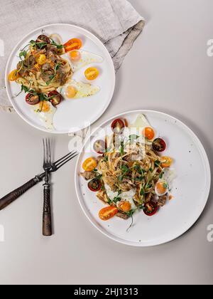 Pasta con tacchino stufato, pomodori ciliegini ed erbe fresche. Uova di quaglia fritte. Due porzioni su sfondo chiaro. Delizioso e salutare Mediterraneo r Foto Stock