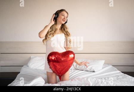 San Valentino, Festa della Donna, anniversario. Una giovane donna in cuffia si siede a letto e festeggia il 14 febbraio con un pallone a forma di cuore rosso Foto Stock